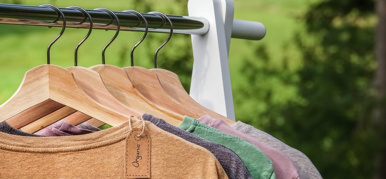 Organic clothes. Natural colored t-shirts hanging on wooden hangers in a row. Eco textile tag. Green forest, nature in background.